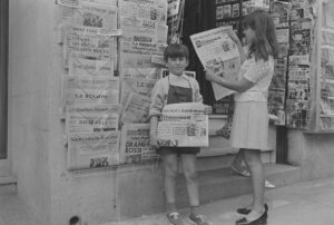 Garçon et fille posant avec des éditions du journal « L'Indépendant » devant un kiosque à journaux